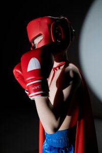 A young child boxer wearing red gear and cape strikes a powerful, heroic pose in a dimly lit environment.